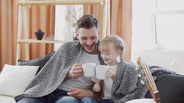 Le jeune père et sa fille sont assis sur un canapé couvert d'une couverture avec de grandes tasses blanches et parle — Video