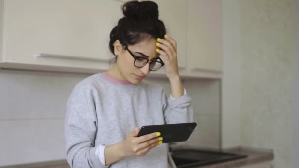 Mujer joven lee algo en su tableta en la cocina — Vídeos de Stock