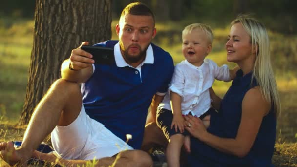 행복 한 가족은 풀밭에 앉아서 해 가질 때 아기와 함께 셀카를 하고 있다. 아버지와 어머니는 아기와 함께 전화기로 자신의 사진을 찍는다 — 비디오
