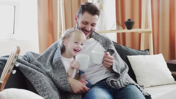 Ο νεαρός πατέρας και η κόρη του κάθονται στον καναπέ σκεπασμένοι κουβέρτα με μεγάλα λευκά φλιτζάνια και συνομιλίες — Αρχείο Βίντεο
