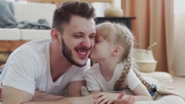 Feliz padre y su hija se acuestan en el suelo. Pequeña linda chica besa a su padre — Vídeo de stock