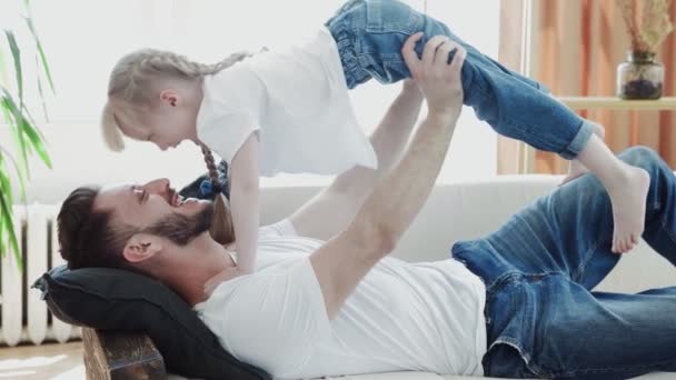 Junger Mann in Jeans und weißem T-Shirt spielt mit seiner Tochter auf der Couch — Stockvideo