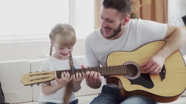 Padre joven enseña a su hija a tocar la guitarra en casa — Vídeo de stock