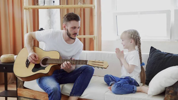 Pouco bonito menina palmas mãos quando seu belo pai estão tocando guitarra enquanto sentado no sofá em casa — Fotografia de Stock