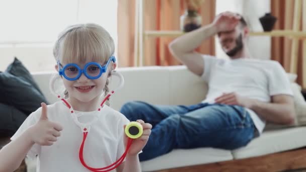 Little funny kid girl dressed in medical uniform playing as doctor with father on background — Stock Video