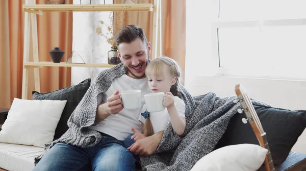 Baba ve kızı. Yakışıklı genç bir adam ve küçük tatlı bir kız evdeki kanepede oturuyor, kendilerini sıcak bir battaniyeyle örtüyorlar, eğleniyorlar, ellerinde beyaz bardaklarla. Baba ve çocuk gülüyor. Babalar Günü