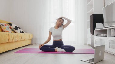 Evde çalışan bir kadın, evdeki dizüstü bilgisayarında eğitmeninin yardımıyla internet hizmetlerini kullanıyor. Zayıf, Afro-Amerikan kadın minderde yoga yapıyor. Boyun geren kadın