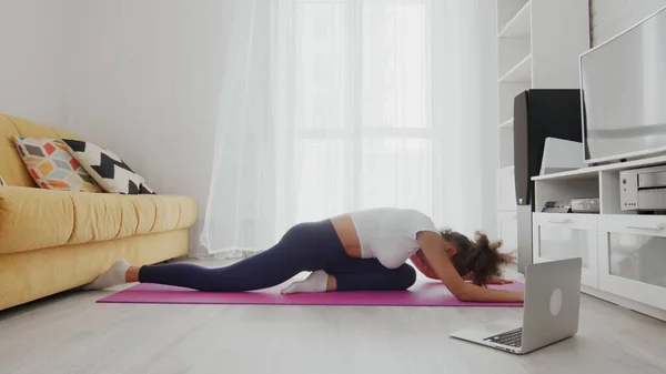 On-line home work out woman using internet services with help of her instructor on laptop at home. Slim sporty african american woman practicing yoga on a mat. Young woman doing yoga workout in room