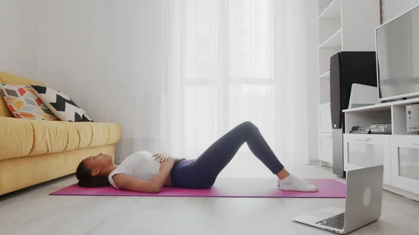 On-line home work out woman using internet services with help of her instructor on laptop at home. Slim sporty african american woman practicing yoga on a mat. Woman having a rest after training