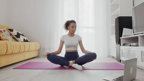 On-line home work out woman using internet services with help of her instructor on laptop at home. Slim sporty african american woman sits in lotus pose and look at the laptop