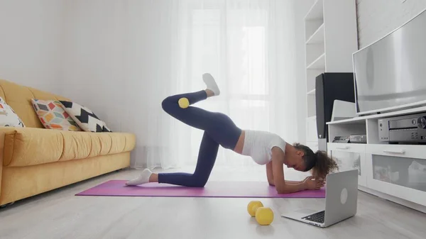 On-line home work out γυναίκα που χρησιμοποιεί υπηρεσίες διαδικτύου με τη βοήθεια του εκπαιδευτή της στο laptop στο σπίτι. Slim σπορ αφρικάνικη αμερικανική γυναίκα ανύψωσης δεξί πόδι με κίτρινο αλτήρα στο χαλί — Φωτογραφία Αρχείου