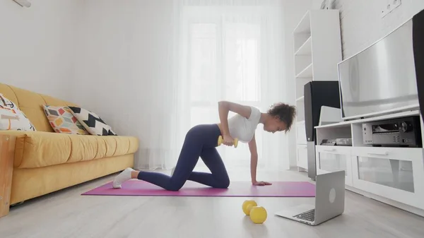 On-line home work out woman using internet services with help of her instructor on laptop at home. Slim sporty african american woman pulling up a yellow dumbbell on mat