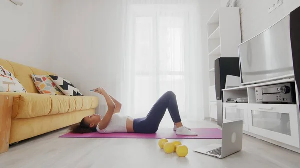 On-line home work out woman using internet services with help of her instructor on laptop at home. Slim sporty african american woman lays down on mat and uses phone after exercises at home