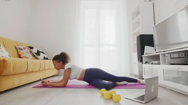 On-line home work out woman using internet services with help of her instructor on laptop at home. Slim sporty african american woman lays down on mat and uses phone after exercises at home