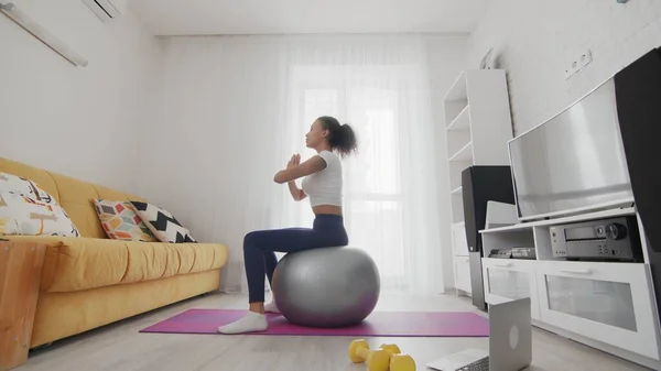On-line home work out woman using internet services with help of her instructor on laptop at home. Slim sporty african american woman sits and meditates on fitball
