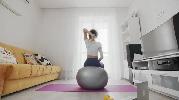 On-line home work out woman using internet services with help of her instructor on laptop at home. Slim sporty african american woman sits on fitball and training right triceps