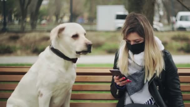 검은 마스크를 쓴 젊은 여성 이 리트리버 래브라도 개와 함께 벤치에 앉아 있고 격리 코로나 바이러스 COVID-19 전염병 이 유행하는 동안 시내 공원에서 전화를 사용 한다. — 비디오