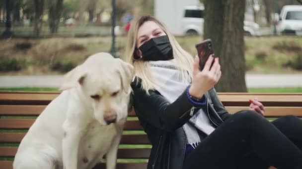 Mujer joven con máscara negra se sienta con perro labrador retriever en el banco y haciendo selfie en el banco en el parque. en el parque de la ciudad durante la pandemia del coronavirus COVID-19 de cuarentena — Vídeos de Stock