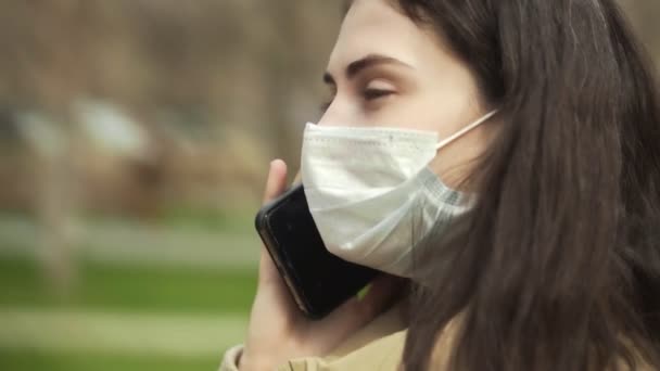 Vista lateral de la mujer joven al aire libre con mascarilla médica para la protección de las conversaciones de virus en el teléfono. Concepto pandemia de coronavirus, SARS, MERS, COVID-19, cuarentena, período de aislamiento . — Vídeo de stock