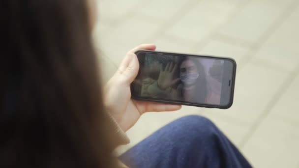 Jonge vrouw met een beschermend masker die video kijkt met haar smartphone in het stadspark. Pandemische Covid-19 bescherming tegen coronavirus. — Stockvideo