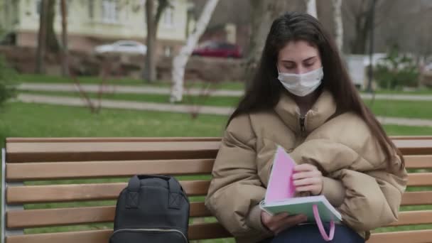 Giovane donna in maschera protettiva scrive al taccuino nel parco della città. Pandemia Covid-19 protezione coronavirus . — Video Stock