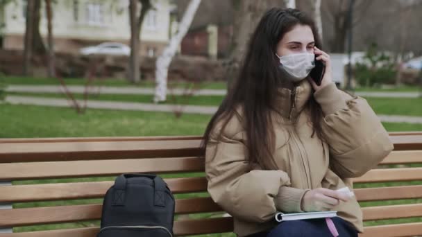 Mujer joven con máscara protectora escribe a diario en el parque de la ciudad. Pandemic Covid-19 coronavirus protection . — Vídeos de Stock