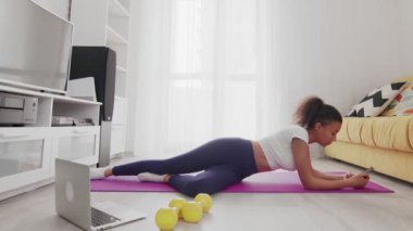 Genç Afro-Afrikalı Amerikalı kadın paspasa uzanıyor ve evdeki egzersizlerden sonra telefonu kullanıyor.