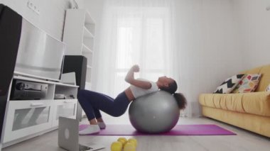 Genç Afrikalı Afrikalı Amerikalı kadının antrenmanlarını evde dizüstü bilgisayarında eğitmeninin yardımıyla fitball üzerinde yapıyor.