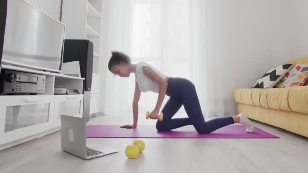 Mladá sportovní africká americká žena vytáhnout žlutý činka na podložce s pomocí svého instruktora on-line na notebooku doma — Stock video