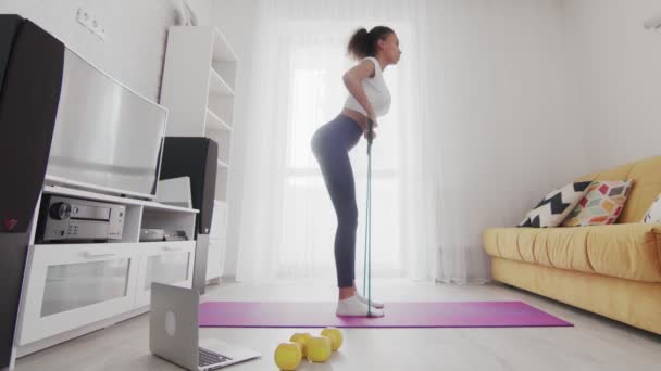 Side view of young sporty african american woman exercising with resistance band on mat with help of her instructor online on laptop at home — Stock Video