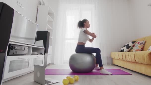 Vista lateral de la joven afroamericana deportiva hace ejercicio de rotación en fitball con la ayuda de su instructor en línea en el ordenador portátil en casa — Vídeos de Stock