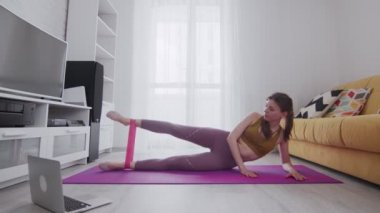 Genç ve güzel bir kadın evde spor yapıyor. Paspasa uzanır ve direnç bandıyla bacağını kaldırır.