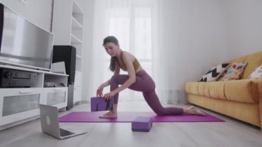 Genç ve güzel bir kadın menekşe paspasın üzerinde bacak esnetme alıştırması yapıyor, iki tuğla kullanıyor ve online talimatlarla dizüstü bilgisayara bakıyor.