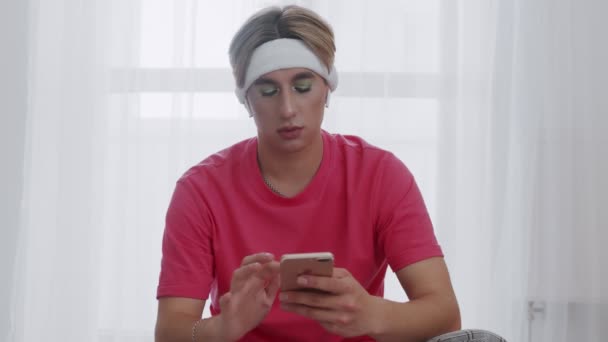 Jovem transexual homossexual homem com maquiagem e com heapdones senta-se no tapete fitnes e usando telefone — Vídeo de Stock
