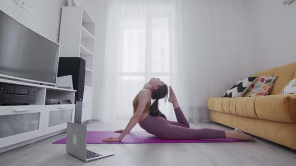 Jonge mooie vrouw doet stretching oefeningen op violette mat thuis. Ze raakt het hoofd met haar voet — Stockvideo