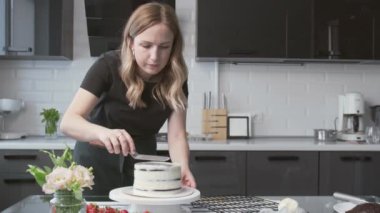 Profesyonel şef kek pişiriyor. Genç ve çekici ev hanımı çikolatalı kekin üzerine beyaz krema koymak için metal spatula ve döner masa kullanıyor.
