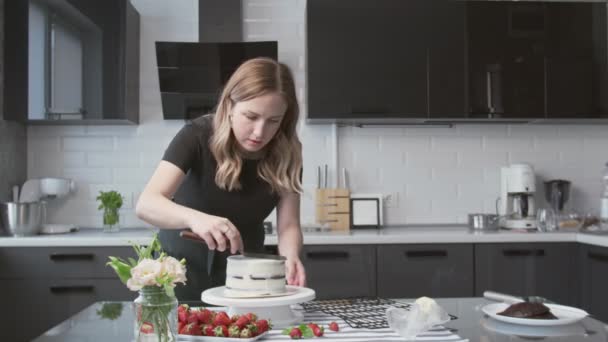 Επαγγελματίας σεφ μαγειρεύει κέικ. Νεαρή ελκυστική νοικοκυρά χρησιμοποιεί μεταλλική σπάτουλα και περιστρεφόμενο τραπέζι για να ευθυγραμμίσει λευκή κρέμα σε κέικ σοκολάτας — Αρχείο Βίντεο