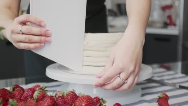 Профессиональный повар готовит торт. Молодая привлекательная домохозяйка использует большую пластиковую лопатку и вращающийся стол, чтобы выровнять белый крем на шоколадном торте — стоковое видео