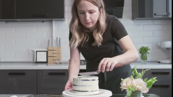 Профессиональный повар готовит торт. Молодая привлекательная домохозяйка использует металлическую лопатку и вращающийся стол для выравнивания белого крема на шоколадном торте — стоковое видео