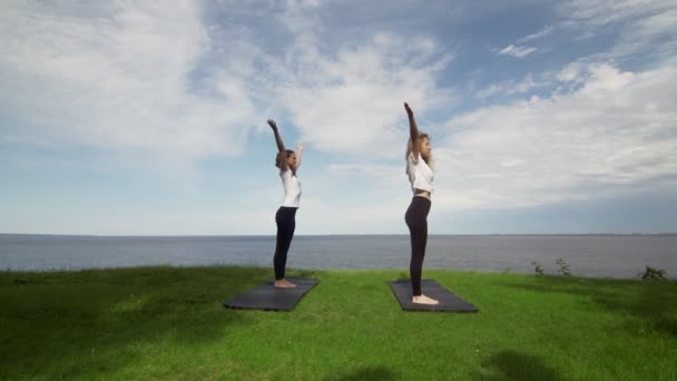 Dos mujeres jóvenes practican yoga en la costa cerca del lago o el mar. Entrenamiento Señor de la Danza pose . — Vídeos de Stock