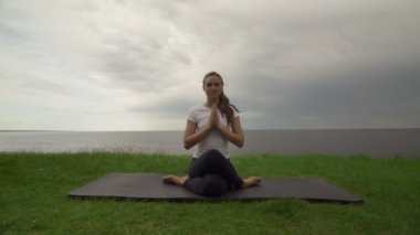 Genç, formda bir kadın göl ya da deniz kıyısında yoga yapıyor. Kadın diz çöküp meditasyon yapıyor.