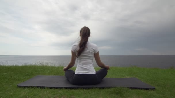 Young fit woman practice yoga on coast near the lake or sea. Woman sitting in lotus pose and meditating — Stock Video