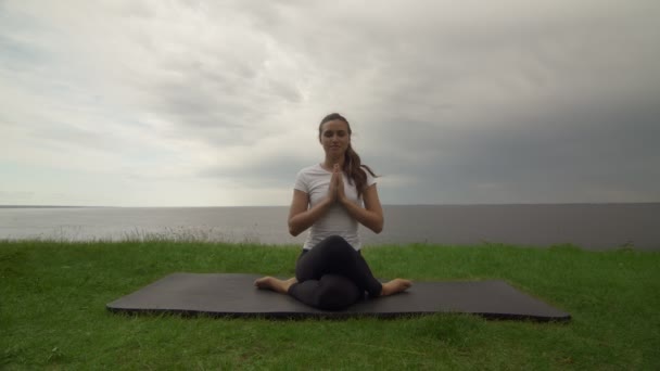 Young fit woman practice yoga on coast near the lake or sea. Woman sitting in Knee Pile pose and meditating — Stock Video