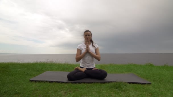 Young fit woman practice yoga on coast near the lake or sea. Woman sitting in lotus pose and meditating — Stock Video
