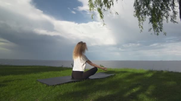 Back view of young fit woman practice yoga on coast near the lake or sea. Woman sitting in lotus pose and meditating — Stock Video