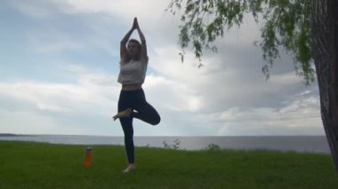 Genç ve formda bir kadının göl ya da denizin kıyısında yoga yapışının arkası. Kadın ayakta yarım lotus pozu veriyor.