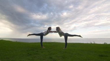 Aktif kıyafetli iki kadın kamerada poz veriyor ve yoga savaşçısı III Virabhadrasana 'yı birlikte gösteriyorlar.