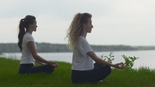 Side view of two young fit women practice yoga on coast near the lake or sea. Women sitting in lotus pose and meditating — Stock Video