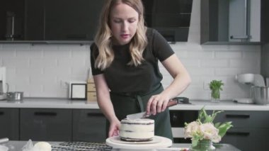 Profesyonel şef kek pişiriyor. Genç ve çekici ev hanımı çikolatalı kekin üzerine beyaz krema koymak için metal spatula ve döner masa kullanıyor.