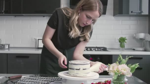 Επαγγελματίας σεφ μαγειρεύει κέικ. Νεαρή ελκυστική νοικοκυρά χρησιμοποιεί μεταλλική σπάτουλα και περιστρεφόμενο τραπέζι για να ευθυγραμμίσει λευκή κρέμα σε κέικ σοκολάτας — Αρχείο Βίντεο
